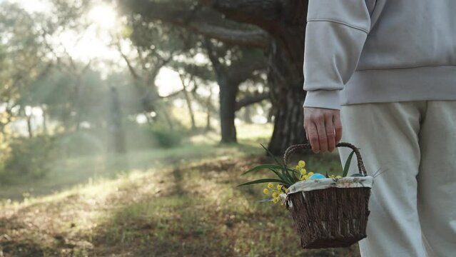 A Woman in White Stands in the Forest, Swinging a Basket with Easter Eggs in her Hands. Sunbeams Break Through the Trees Ahead.
