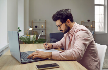 Side view of a young serious brunette businessman in glasses working with laptop computer at home...