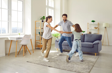 Obraz premium Overjoyed family with child at home actively spends time together dancing to music. Young Caucasian mom, dad and their preteen daughter are holding hands and dancing in living room. Family concept.