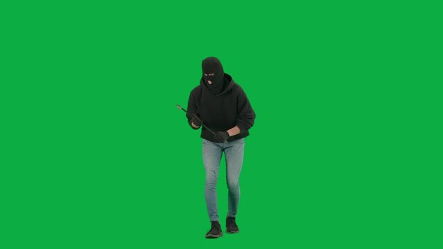 Portrait of thief on chroma key green screen background. Man robber in balaclava and hoodie walking holding crowbar in hands looks around.