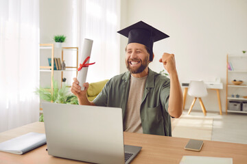 Happy excited graduate student man in hat sitting at the desk with diploma in hands in front of...