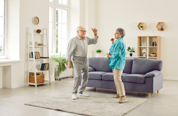 Full length photo of happy senior elderly family couple wife and husband dancing in the living room at home. Smiling retired man and woman having fun, enjoying spending time together on retirement.