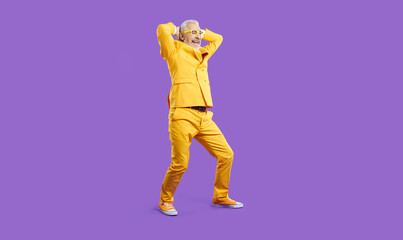 Funny cheerful senior man having fun in fashion studio. Happy crazy old man wearing bright yellow party suit and eyeglasses dancing with his hands behind head isolated on solid purple background