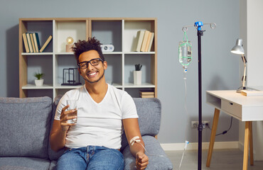 Young happy african american man sitting on the couch in the clinic holding a glass of water while receiving IV drip infusion and vitamin therapy in his blood. Person receiving injection therapy. - obrazy, fototapety, plakaty