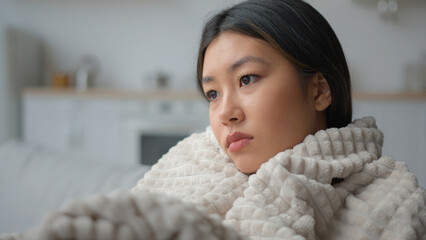 Sad depressed Asian woman in home kitchen alone wrapped warm blanket thinking deep thoughts problem...