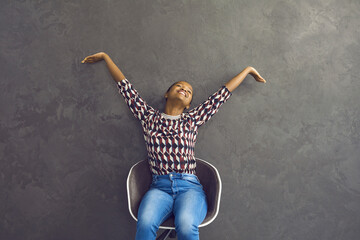 Happy african american woman relaxed stretches her hands while sitting on a chair on a gray...