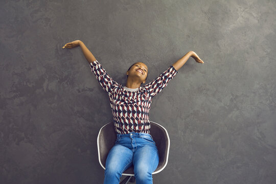 Happy African American Woman Relaxed Stretches Her Hands While Sitting On A Chair On A Gray Background. Woman Feels Happy Resting After Work. Concept Of Happiness, Peace And Relaxation.