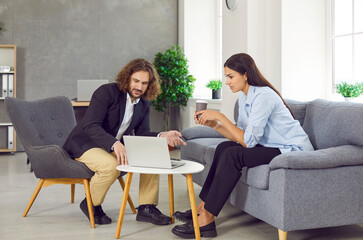 Two business people discussing something during office meeting. Young man and woman sitting on sofa and chair in office, drinking coffee, looking at laptop computer and talking 