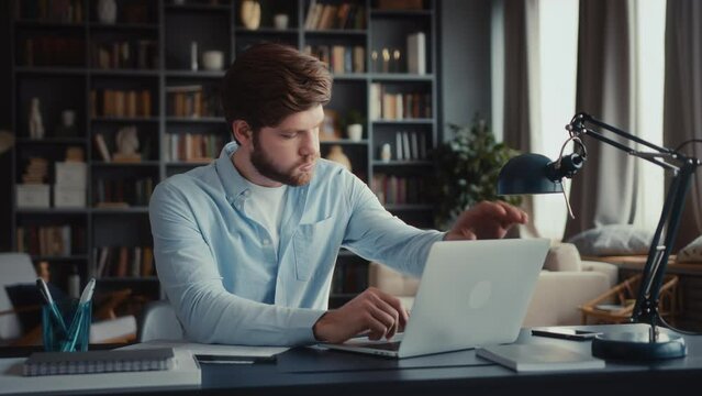 young serious and focused man sits at table in home office and works at laptop, looks at screen attentively, gets disappointed news, closes laptop frustrated shaking head. puts laptop in bin
