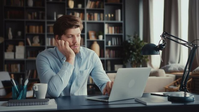 young tired and burn out man in blue shirt sits at table in home office and works at laptop, bored and unmotivated types slowly on keyboard without energy and interest, procrastinating