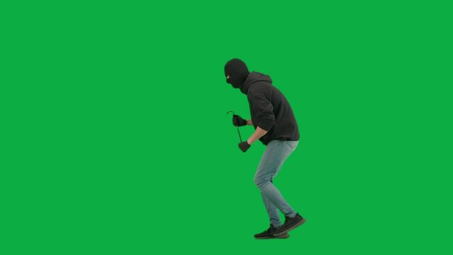 Portrait of thief on chroma key green screen background. Man robber in balaclava and hoodie walking holding crowbar in hands looks around. Side view.