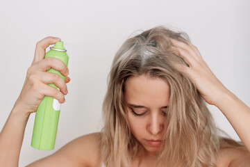 Young blonde woman with dirty greasy hair spraying dry shampoo on the roots of her hair on a light...