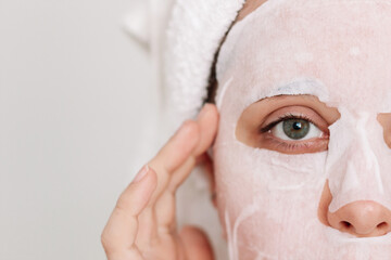 Cropped shot of a young caucasian woman with a white towel on her head after a shower putting a fabric cosmetic moisturizing mask on her face on a light background. Skin care, cosmetology