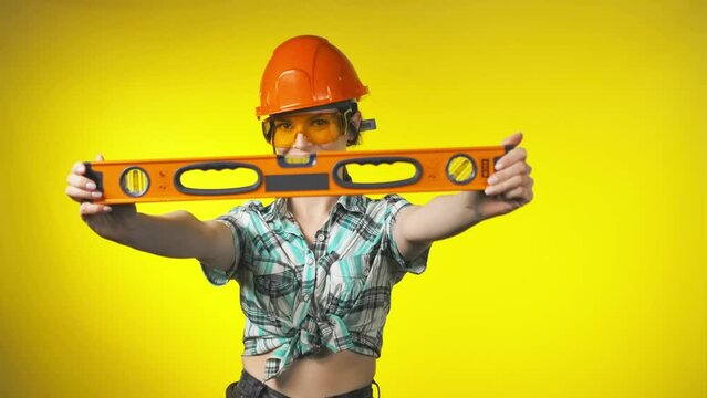 Girl builder in an orange safety helmet is holding a building level ruler in hands in a yellow studio