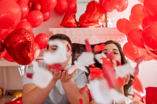 Close up portrait of young man and woman seated close to each other against of white decorative fireplace adorned with red hearts balloons. Couple blowing rose petals towards camera. Valentines Day