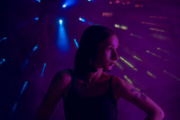 A young woman in black casual clothes poses in a studio with smoke, purple light and beams of multicolored light. The dancer demonstrates elements of experimental hip hop style dance choreography.