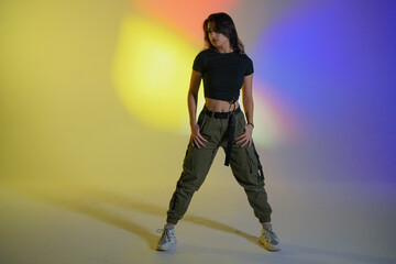 Young woman in black clothes posing in studio with gradient blue, yellow, pink neon light. Dancer demonstrates elements of jazz funk style dance. Contemporary choreography.