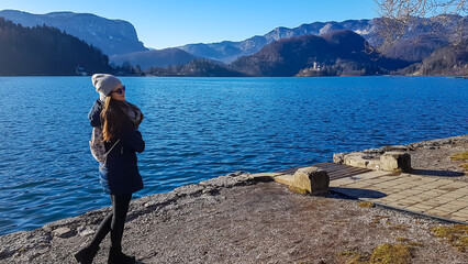 Fototapeta na wymiar Tourist woman with panoramic view of St Mary Church build on small island on alpine lake Bled, Upper Carniola, Slovenia. Serene landscape in Julian Alps in winter. Hills covered with lush green forest