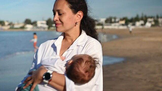 Beautiful delightful woman, loving mother holding her baby boy sleeping in her arms, smiles looking aside while walking along Atlantic beach. People. Family. Leisure activity. Maternity leave