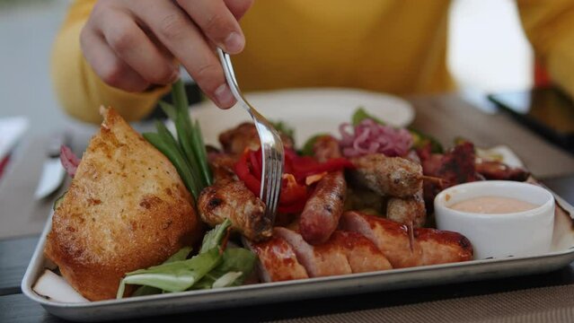 Unrecognizable traveler touches large Balkan dish with assorted meats and vegetables with fork in local authentic bar. Closeup of grilled meat, cevapcici and Serbian pljeskavica in flatbread.