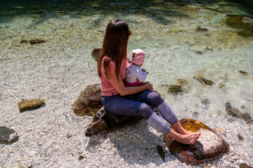 Barefoot woman with baby sitting on rock of Soca River, Bovec, Triglav National Park,Slovenia. Magnificent Soca Valley in Julian Alps. Wanderlust in untamed Slovenian Alps. Hiking in alpine wilderness - obrazy, fototapety, plakaty
