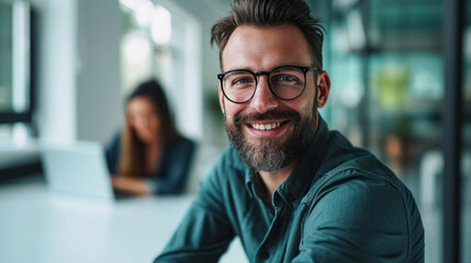 Obraz premium Young man with glasses in the foreground, smiling towards the camera, with coworkers in the background, in a meeting at office
