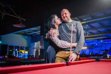 woman affectionately embracing a man from behind at a pool table, both smiling and enjoying a moment together in a neon-lit game hall...