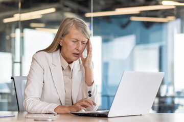 Senior gray-haired businesswoman working in the office, sitting at the desk, holding her head with...