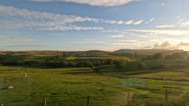 Scenic train ride through rolling hills of Scottish countryside at golden sunset. View from the window of a travelling train passing endless green fields, grazing animals and idyllic small villages.