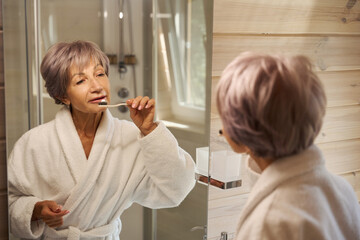 Elderly woman brushes her teeth with toothbrush in front of mirror