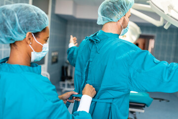Female surgeon tying surgical gown for male colleague in operating room. Two surgeons helping each...