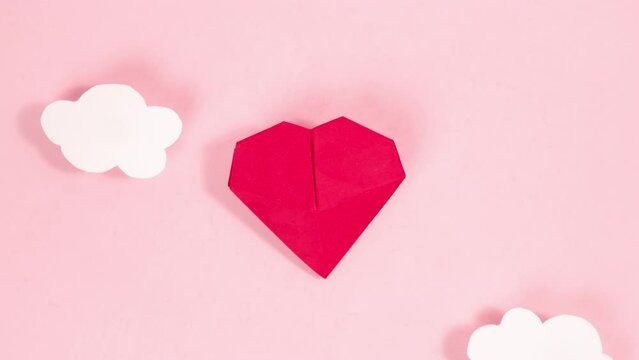 4k Greeting card. Happy Valentine's Day. A red volumetric origami heart flies upward between moving white clouds. Pale pink background. Symbol of love. Looped frame-by-frame animation.
