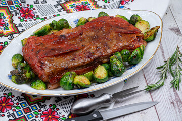 Vegan pork belly roast made with seitan and starch, served on oval plate along with roasted Brussels sprouts. Wooden table,  colorful tablecloth