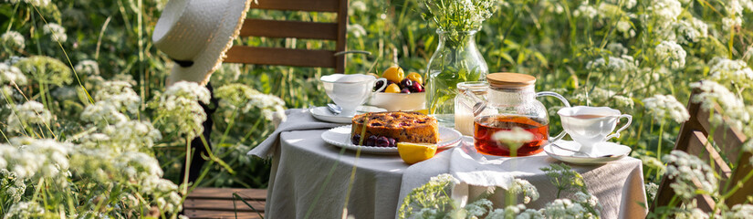 Elegant glamour table setting outdoor in the garden. White porcelain cups, teapot with herbal tea, homemade cherry sweet pie. Family tradition, beautiful place outside. Flower meadow, candles. Banner