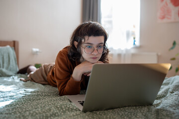 Intrigued concentrated teen girl lying on bed, watching interesting film, video, movie on laptop computer, reading online book attentively, relaxing after working week during weekend, spending pastime
