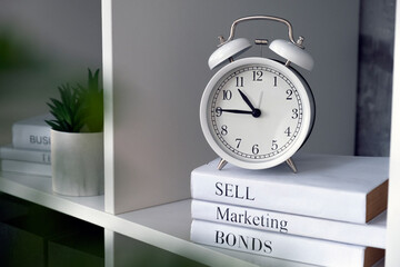 Fototapeta na wymiar White alarm-clock with business books on shelves in office. Business strategy, sell, marketing. Start-up, success and solutions. Investment, analysis and management.