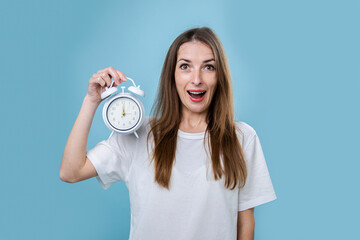 Surprised young woman holding white alarm clock on blue background