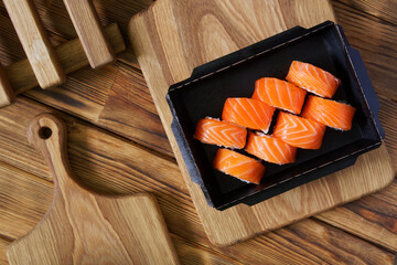 Portion of Philadelphia salmon sushi roll in a black cardboard box from the delivery service on an eco-friendly vintage pine plank table next to a wooden chopping board. Photo