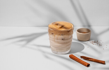 Dalgona coffee. Whipped instant iced coffee with milk in a glass with cinnamon sticks on gray background with shadow.