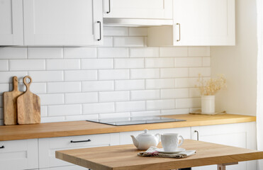 Wooden oak tabletop with a cup of tea and a kettle against the backdrop of a white kitchen with a brick wall with morning sunlight from the window.