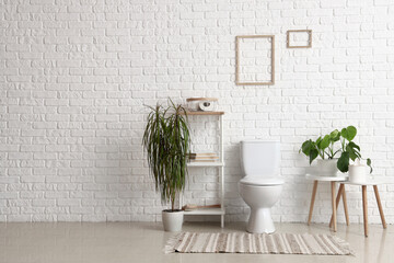 Interior of light restroom with ceramic toilet bowl, shelving unit and houseplants near white brick...