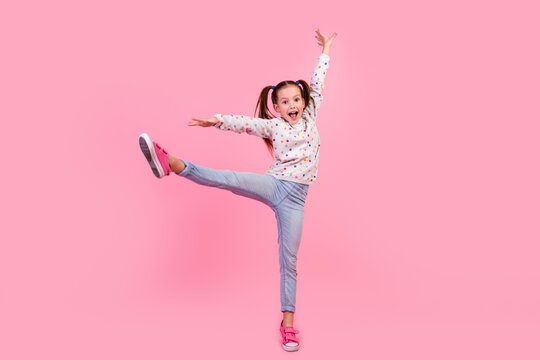 Full length photo of small girl wear stylish sweatshirt jeans raising leg up stand on tiptoe dancing isolated on pink color background