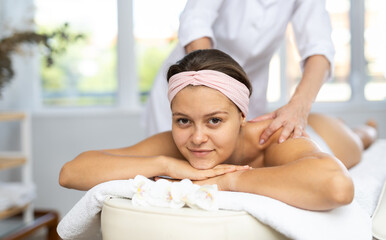 Positive young adult woman relaxing in cozy spa center while professional masseuse doing sports...