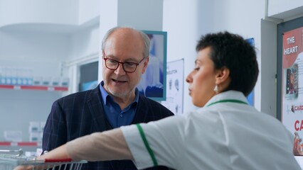 Obraz premium Octogenarian man arriving at pharmacy checkout counter after finding needed medical items, buying disease cure pills with credit card, being helped by druggist with shopping bag