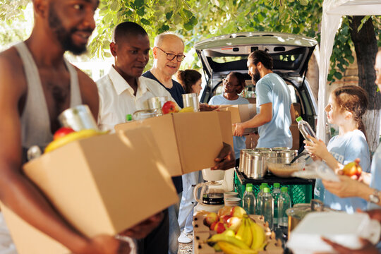 Group of individuals smiling while distributing meal boxes and canned goods to needy. Young volunteers happily provide humanitarian aid to homeless people seniors and individuals seeking shelter.