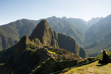 Young woman with hat sitting on lawn looking at Machu Picchu citadel