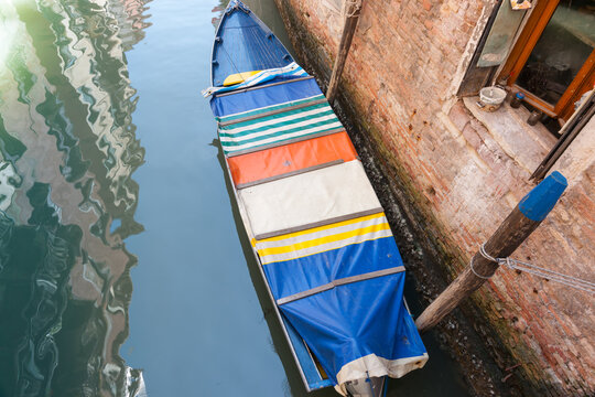 Colourful cover on boat below in canal beside rustic brick wall and window