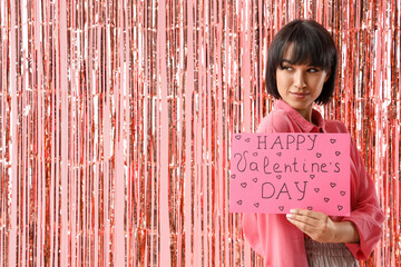 Beautiful young woman holding gift card with text HAPPY VALENTINE'S day and tinsel curtain on pink...
