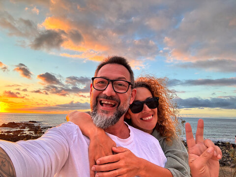 Happy adult couple in love taking selfie picture with romantic wonderful sunset on the ocean in background. Young mature people man and woman enjoying travel destination in summer holiday vacation