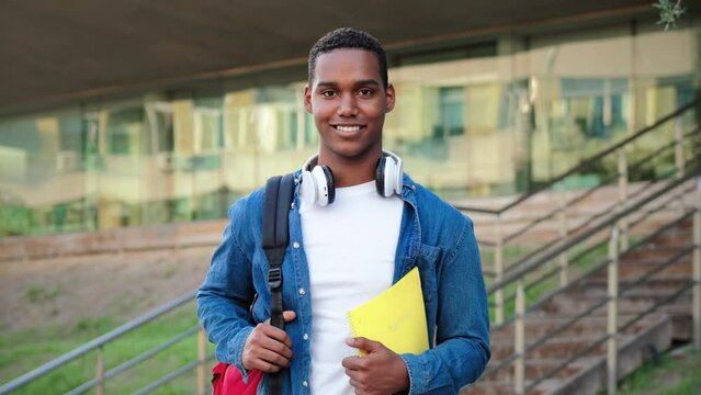 Close up individual portrait of a african american student young man carrying a backpack and note books at university campus. Teenage male smiling and looking at camera. High school guy staring front